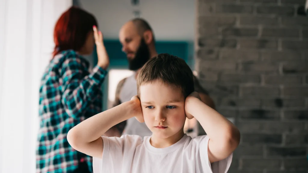 Kind hält sich die Ohren zu, Eltern streiten im Hintergrund – Symbolbild für Streit in der Familie